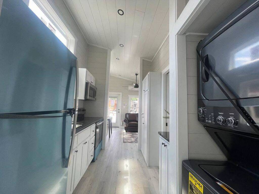 Bright, narrow kitchen with white cabinets and a blue fridge on the left. Stacked black washer and dryer on the right. Cozy living area in the back.