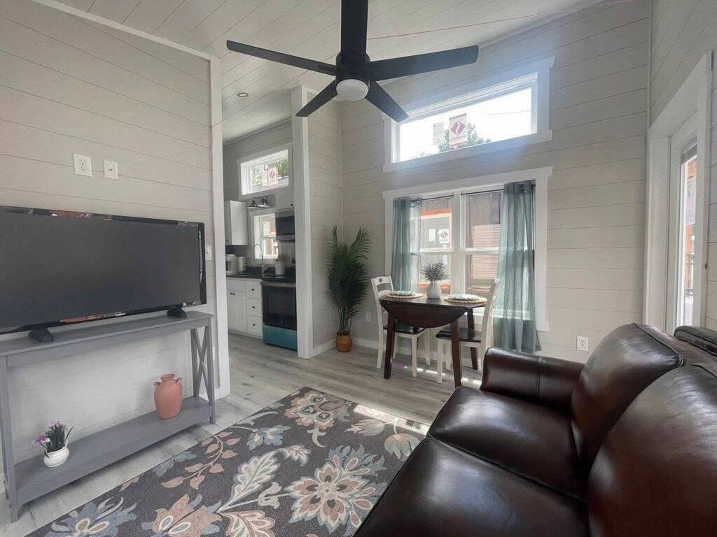 Cozy living room with a brown leather sofa, floral rug, and TV on a gray stand. A round dining table is near a window with teal curtains. Bright and inviting.