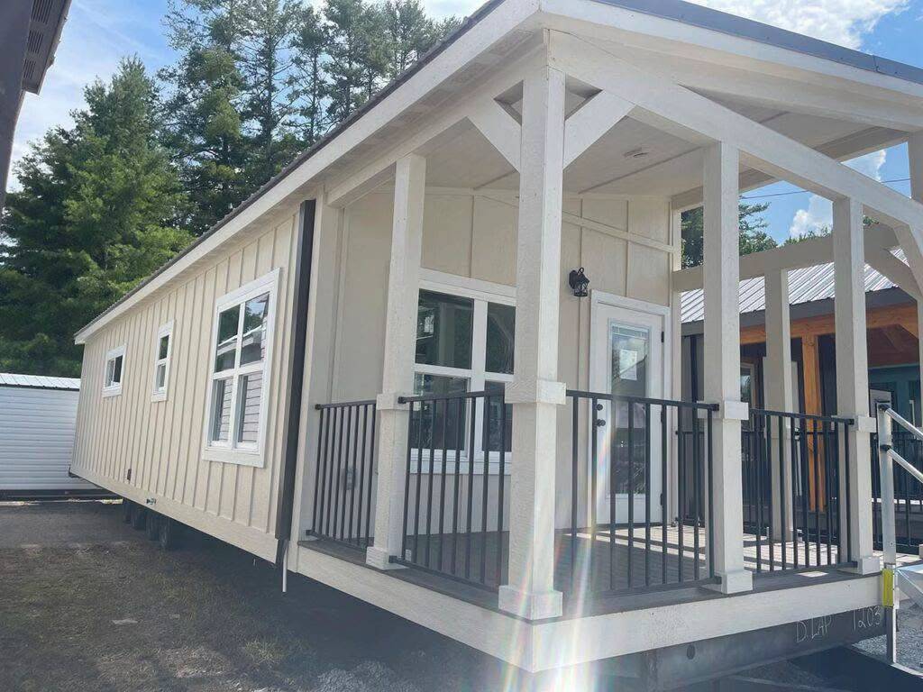 A modern tiny home with beige paneling and large windows, featuring a covered porch with black railings. Surrounded by trees, it conveys a cozy, serene vibe.