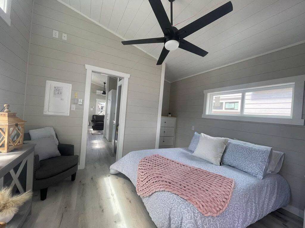 Cozy bedroom with light gray walls, a large black ceiling fan, and a bed with soft blue bedding and a pink throw. Sunlight streams through a small window.