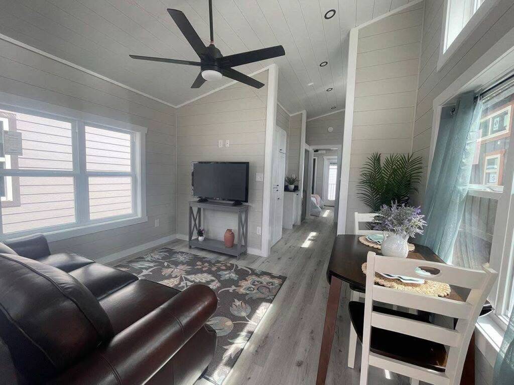 A cozy and bright tiny home interior with a modern design. It features a leather couch, TV on a stand, dining table with flowers, and a ceiling fan.