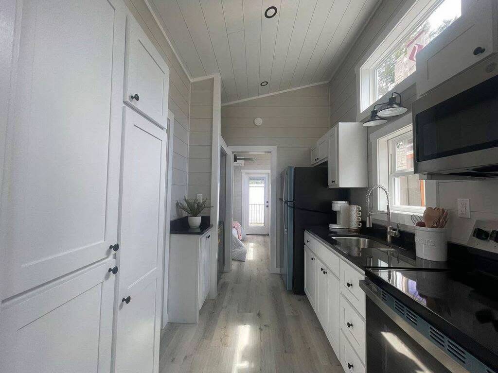 Bright, narrow kitchen with white cabinets, black countertops, and light wood flooring. Sunlight streams through windows, creating a cozy, modern feel.