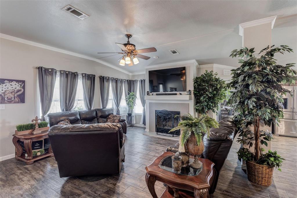 A cozy living room with dark leather sofas facing a fireplace. Large windows with gray curtains allow natural light. Lush green plants and a ceiling fan enhance the space.