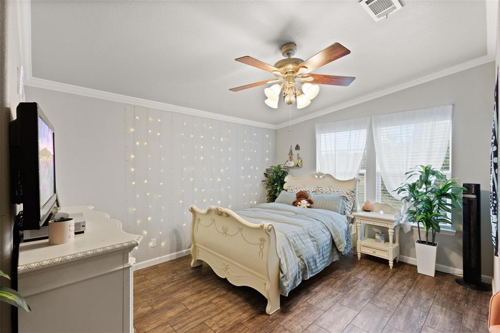 Cozy bedroom with beige walls, wooden floor, an ornate cream bed, and string lights on the wall. Ceiling fan, plants, and a small shelf add warmth.