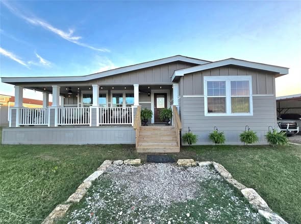 Single-story gray house with a wide front porch and white railings, two large windows, and a centered wooden staircase. Manicured lawn with stone path.