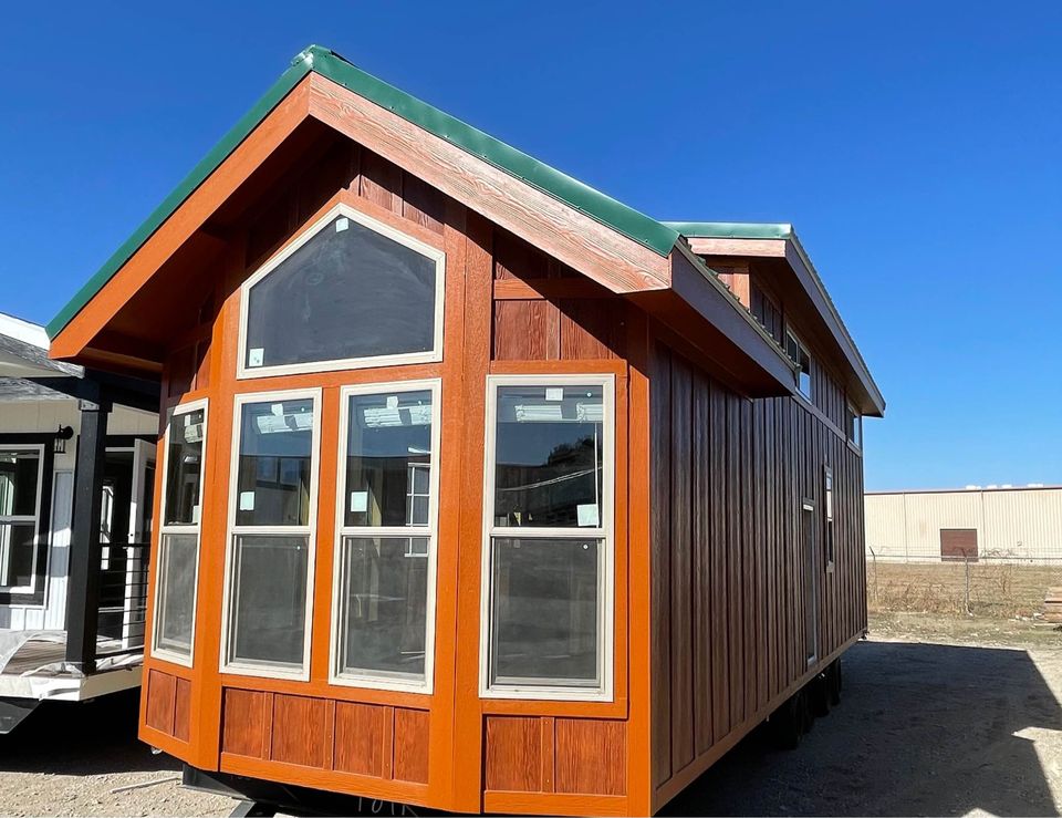 Wooden tiny house with large windows and a green roof under a clear blue sky. The structure conveys a rustic, cozy aesthetic.