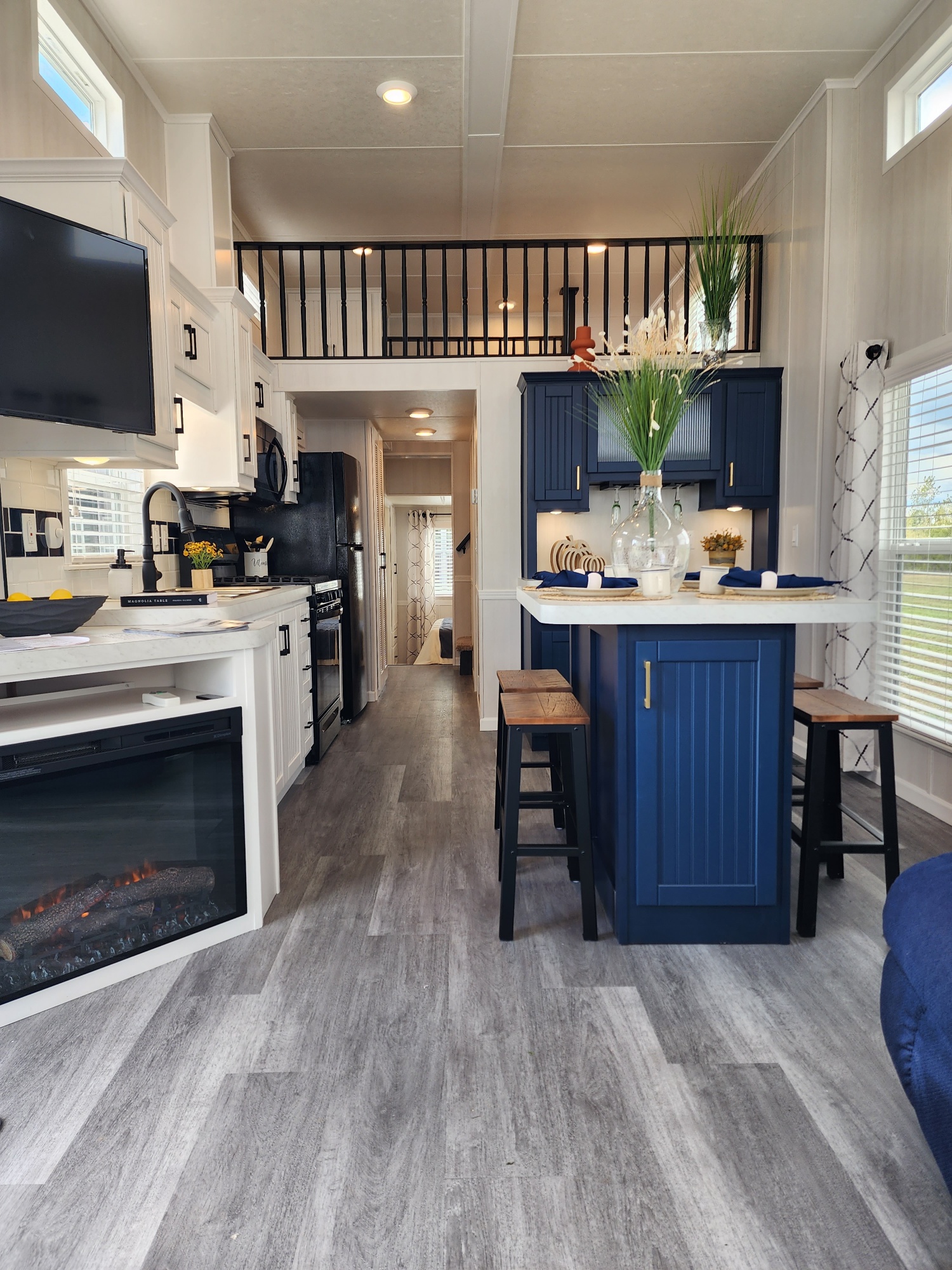 Modern tiny home interior with a cozy kitchen and living area. White cabinets, navy island with stools, faux fireplace, and loft above add charm.