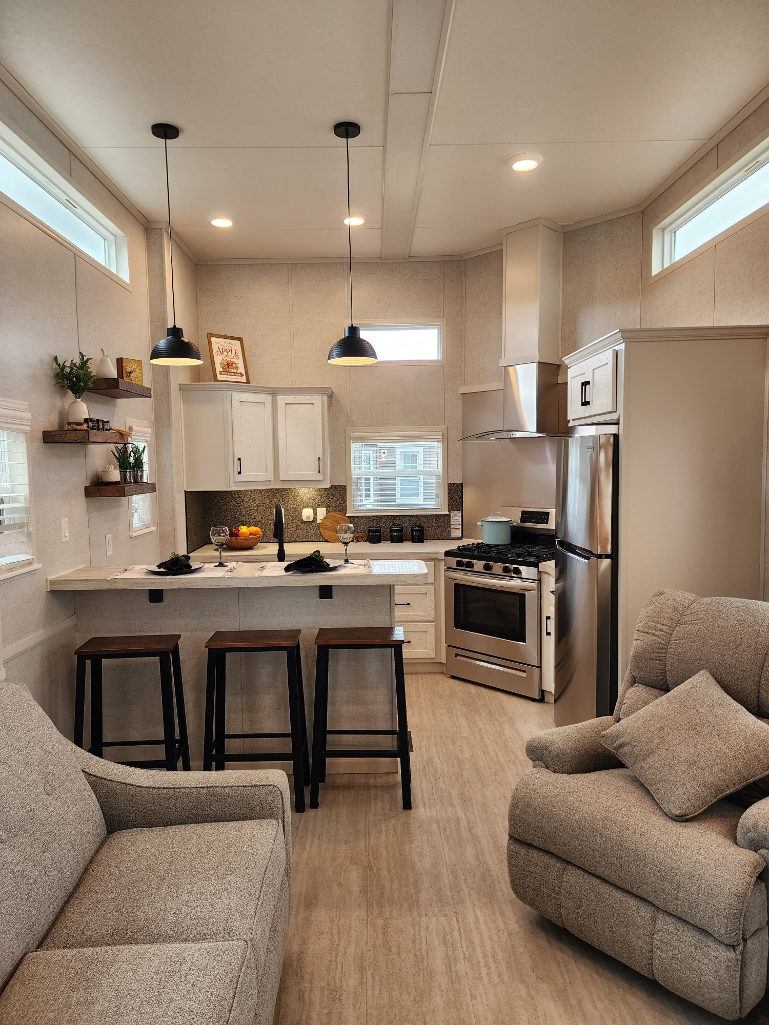 Modern compact kitchen and living area with neutral tones. Includes stainless steel appliances, pendant lights, a cozy sofa, and three bar stools. Clean and inviting space.