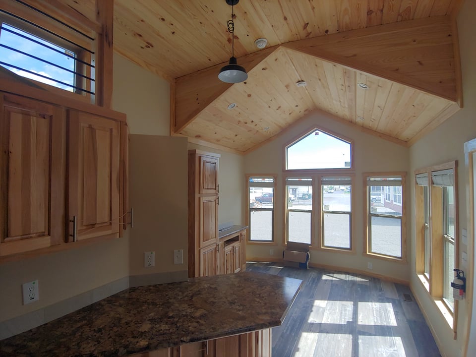 Interior of a tiny home with wooden cabinets, vaulted ceiling, and large windows. Sunlight streams in, creating a warm, cozy atmosphere.