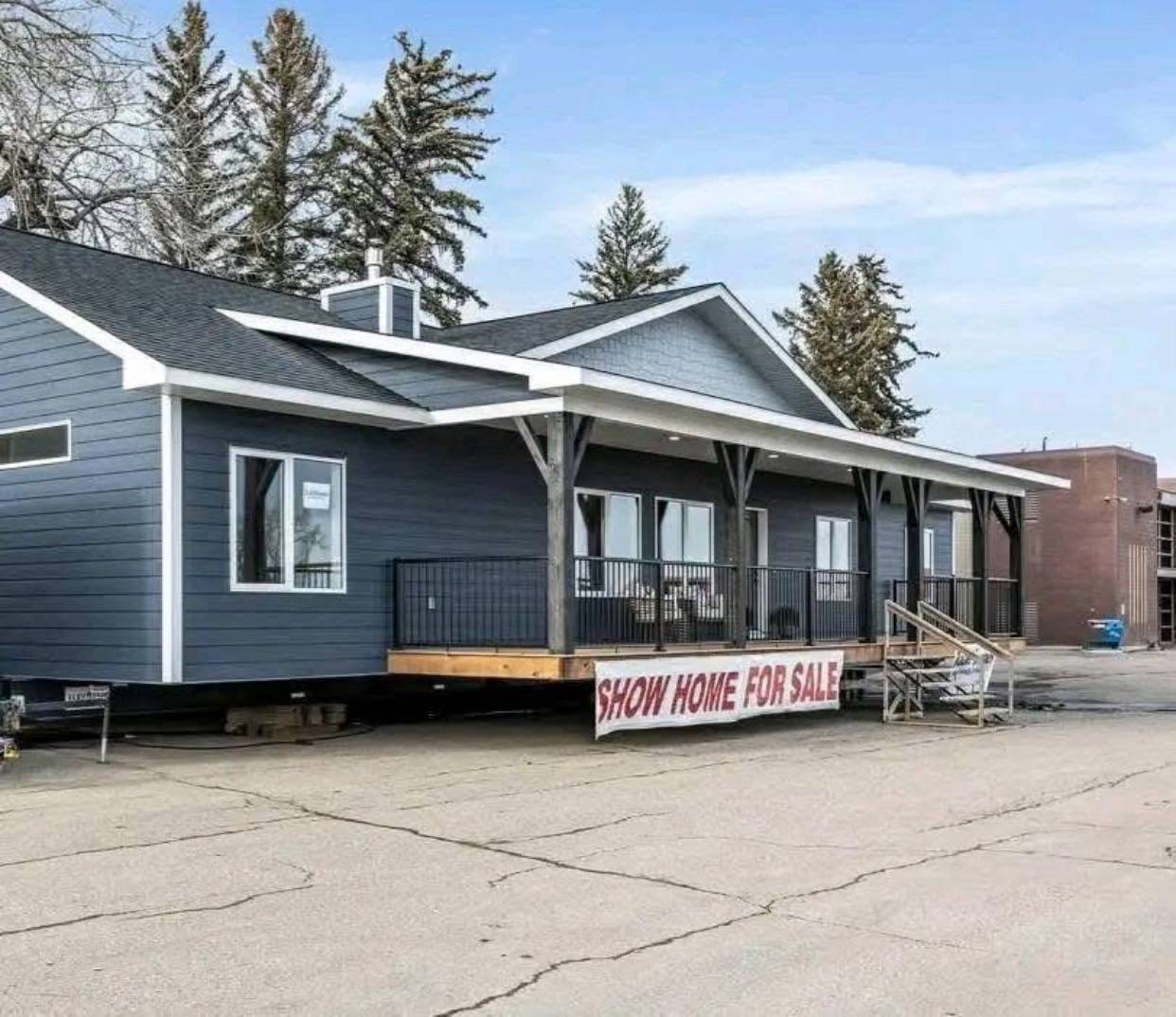 A dark gray show home with white trim is on display outdoors, elevated on a platform. A sign reads "Show Home For Sale." Tall trees and a building are in the background.