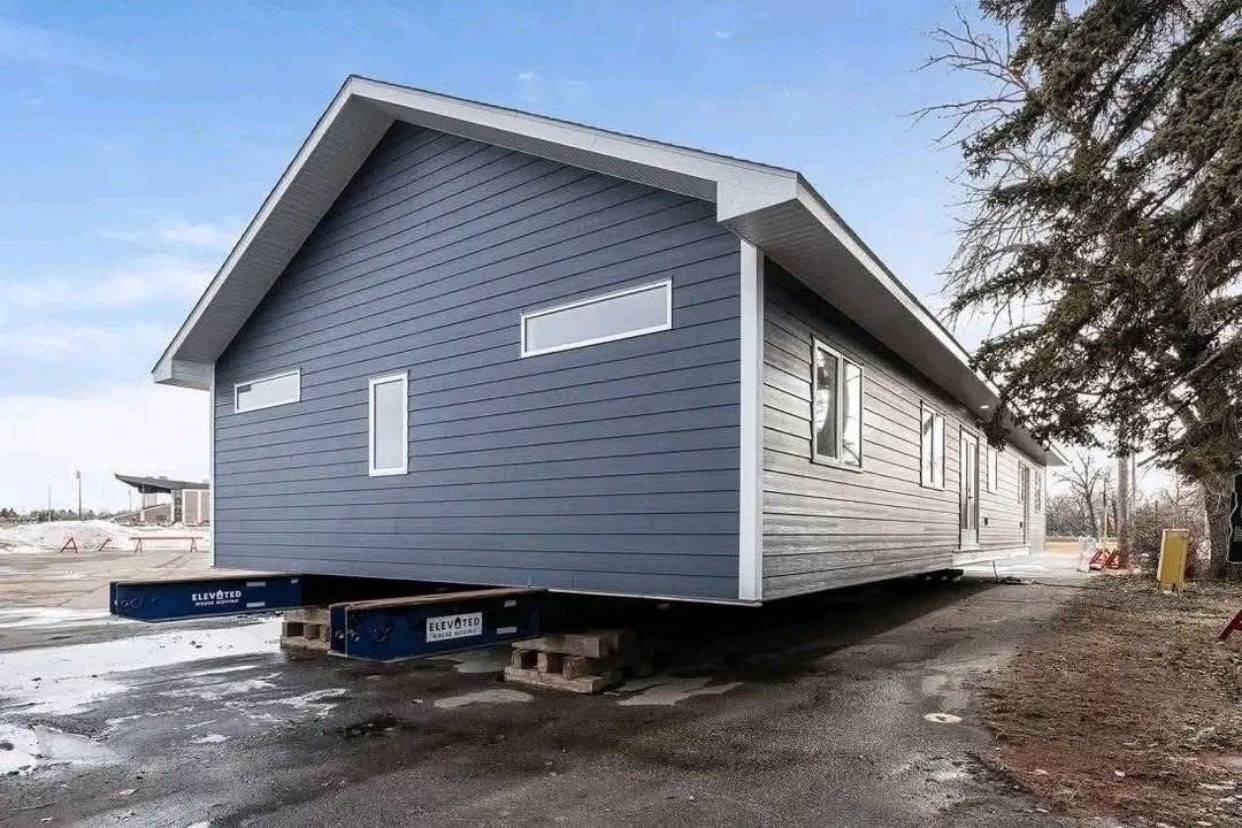 A blue modular home sits elevated on supports, ready for transport. The house features horizontal siding and narrow windows, set against a clear sky backdrop.