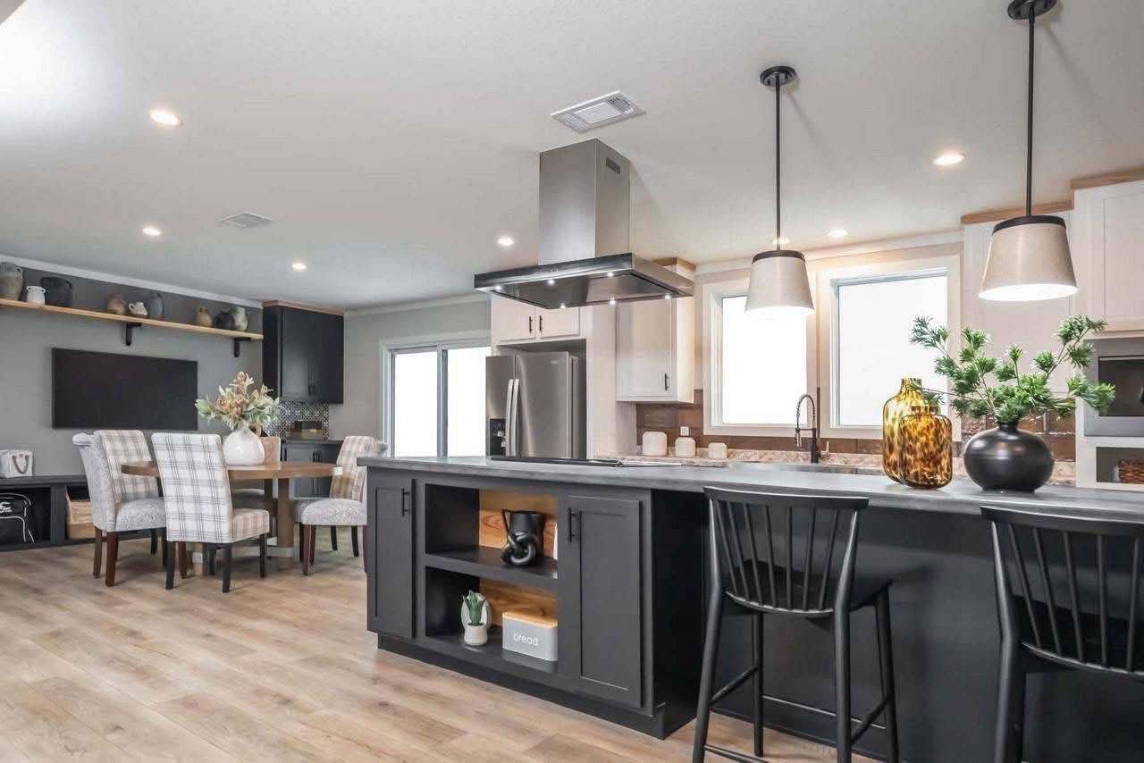 Modern kitchen with a large island, black chairs, pendant lights, and hardwood floors. Dining area in the background with plaid chairs, creating a cozy ambiance.