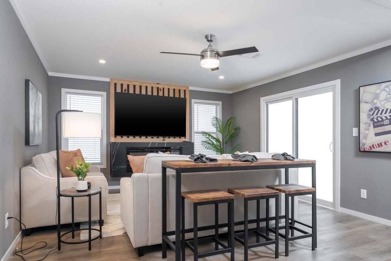 Modern living room with gray walls featuring a ceiling fan, wall-mounted TV, white sofas, rustic wood dining table, stools, and a sliding glass door.