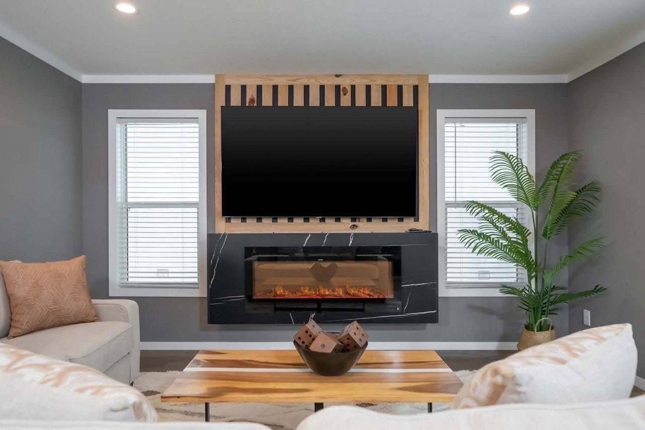 A cozy living room with a modern fireplace under a large flat-screen TV. Soft sofas surround a wooden coffee table with decorative dice. A potted plant adds greenery.