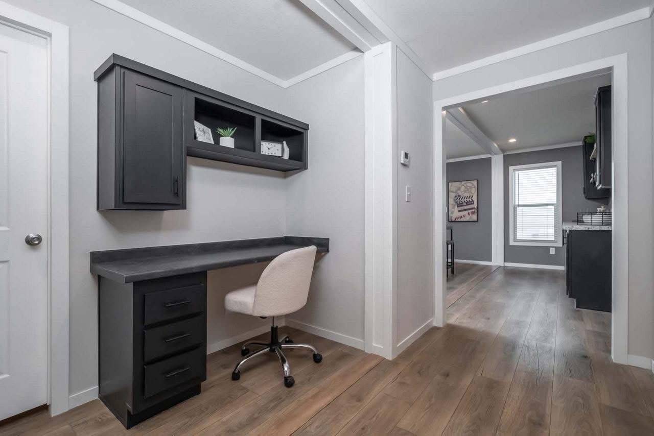 A minimalist home office with a black desk and overhead cabinets, a beige swivel chair, wood floors, and a doorway leading to a bright, airy room.