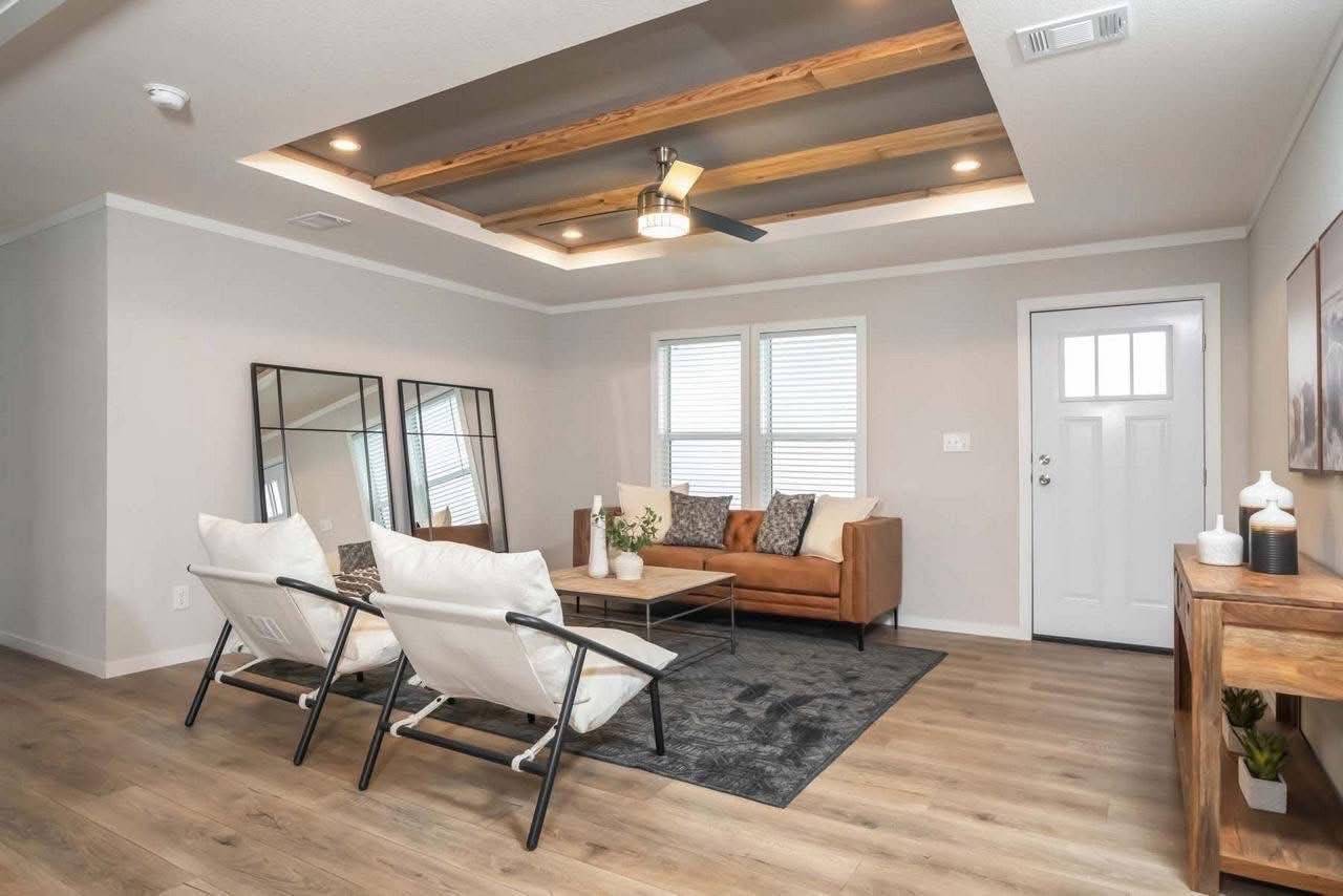 Modern living room with a cozy brown sofa, white armchairs, and wooden accents. Ceiling features recessed lighting, fan, and skylight depth.