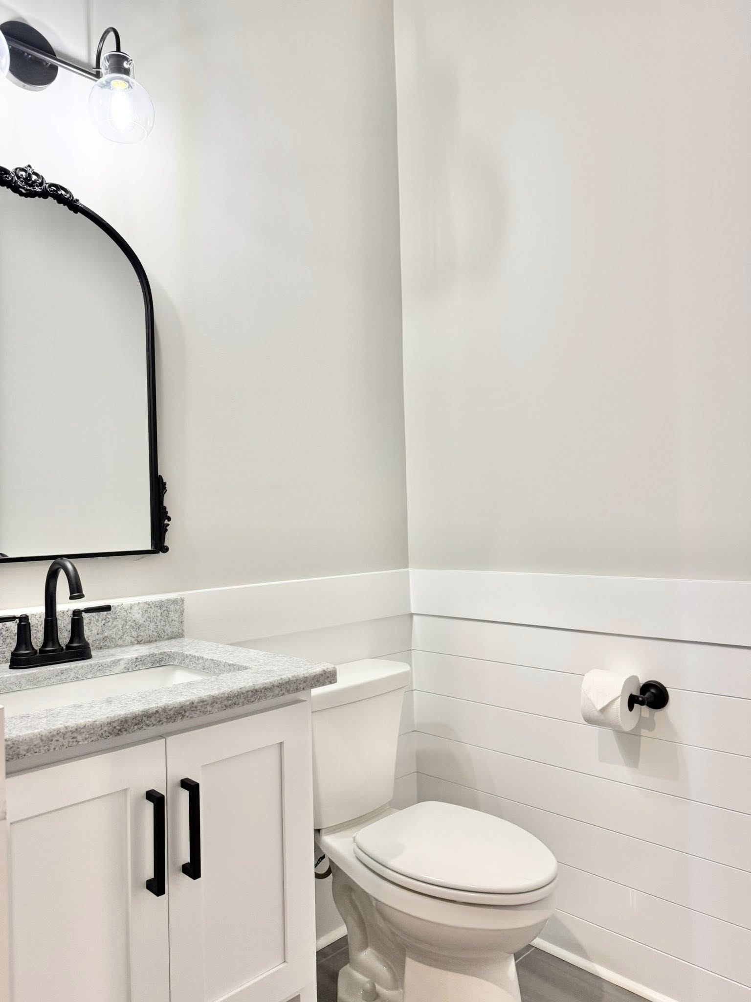 A minimalist bathroom features a white vanity with black handles, a marble countertop, a black-framed oval mirror, and a toilet. The decor is clean and modern.