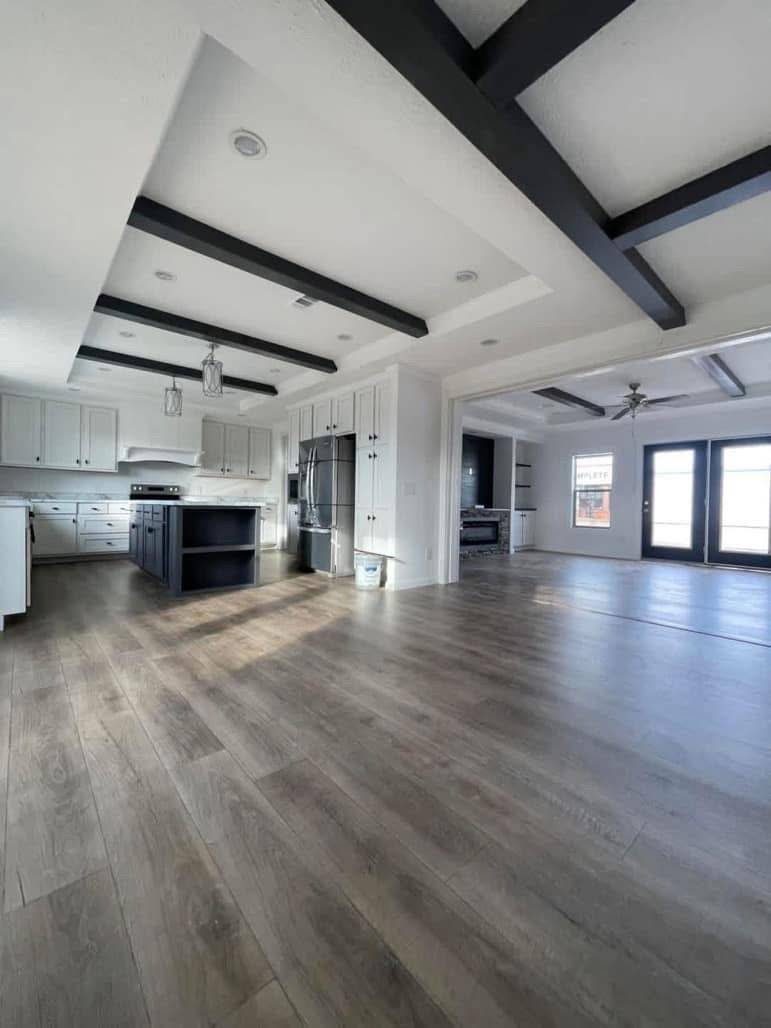 Spacious modern kitchen and living area with dark wooden floors, exposed beams, white cabinets, a central island, stainless steel appliances, and large windows.