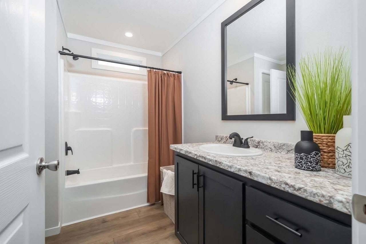 Modern bathroom with light walls and wood flooring. Features a black vanity with a granite countertop, large mirror, white sink, and decorative plants.