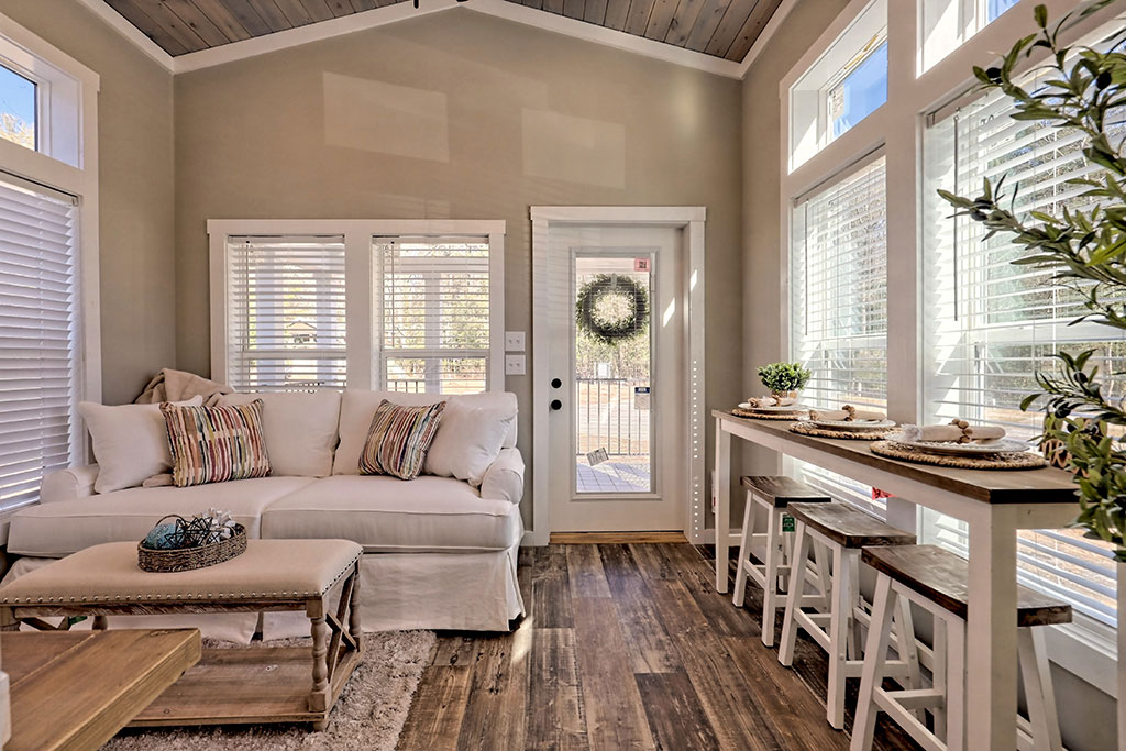 Bright living room with a white sofa, striped pillows, and a wooden coffee table on the left. Bar table with stools on the right, large windows, and a door with a wreath. Cozy and inviting.