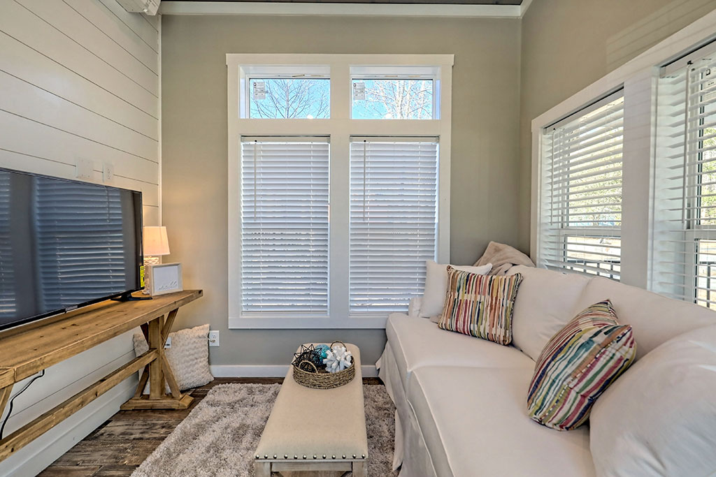 Cozy living room with a white sofa adorned with colorful striped pillows, a wooden coffee table, a TV on a rustic stand, and large windows with blinds.