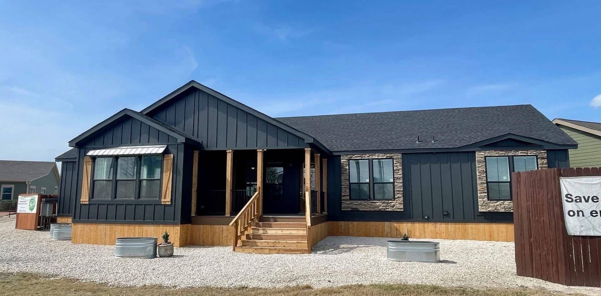 A modern modular home with dark siding and stone accents. It has a gabled roof, front porch with steps, and is surrounded by gravel and grass under a clear blue sky.