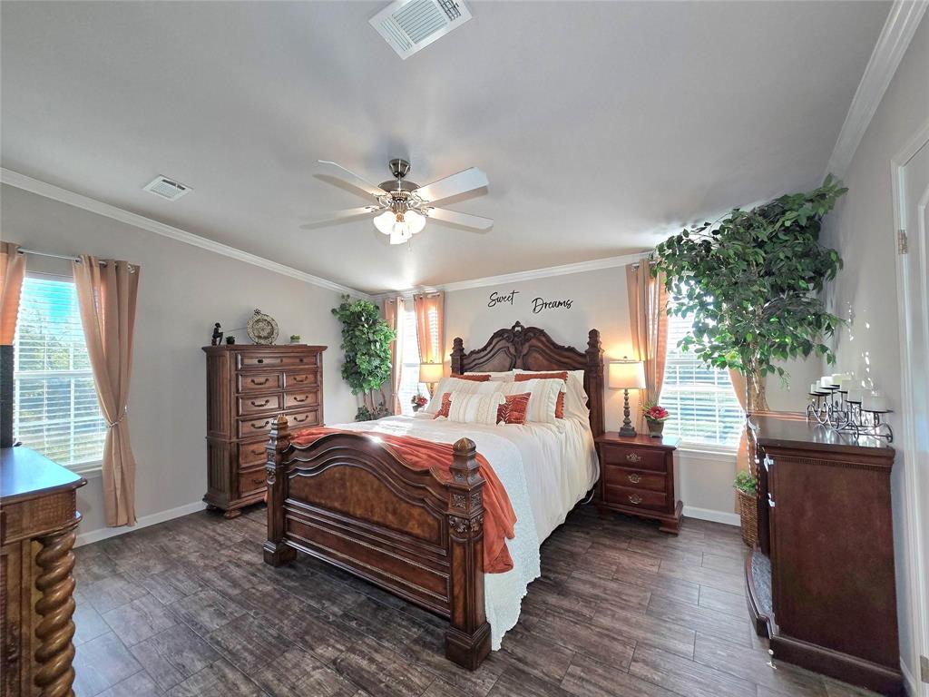 Spacious bedroom with elegant wooden furniture, white and orange bedding, large windows with curtains, ceiling fan, and green plants for a cozy ambiance.