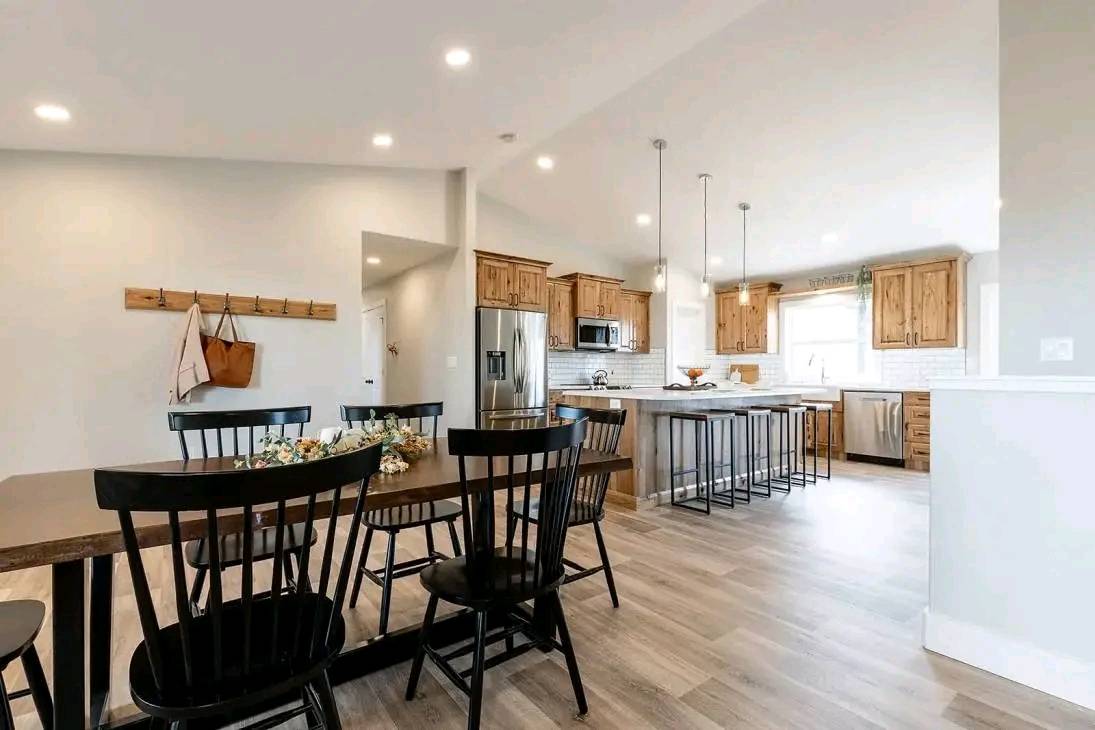 Spacious kitchen-dining area with wooden floors, black dining chairs, and a wooden table. Kitchen features wooden cabinets, stainless appliances, and island seating. Bright, airy ambiance.