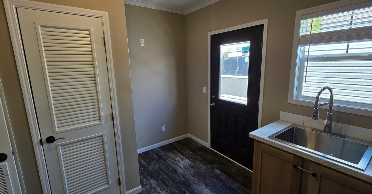 A small laundry room with a dark door, window, and sink beneath another window. Light tan walls, louvered closet door, and dark wood floor.