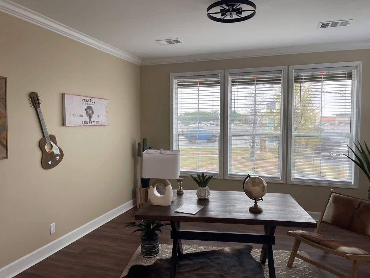 A cozy office with large windows letting in natural light. A wooden desk features a lamp, globe, and plants. A guitar and artwork hang on the beige walls.