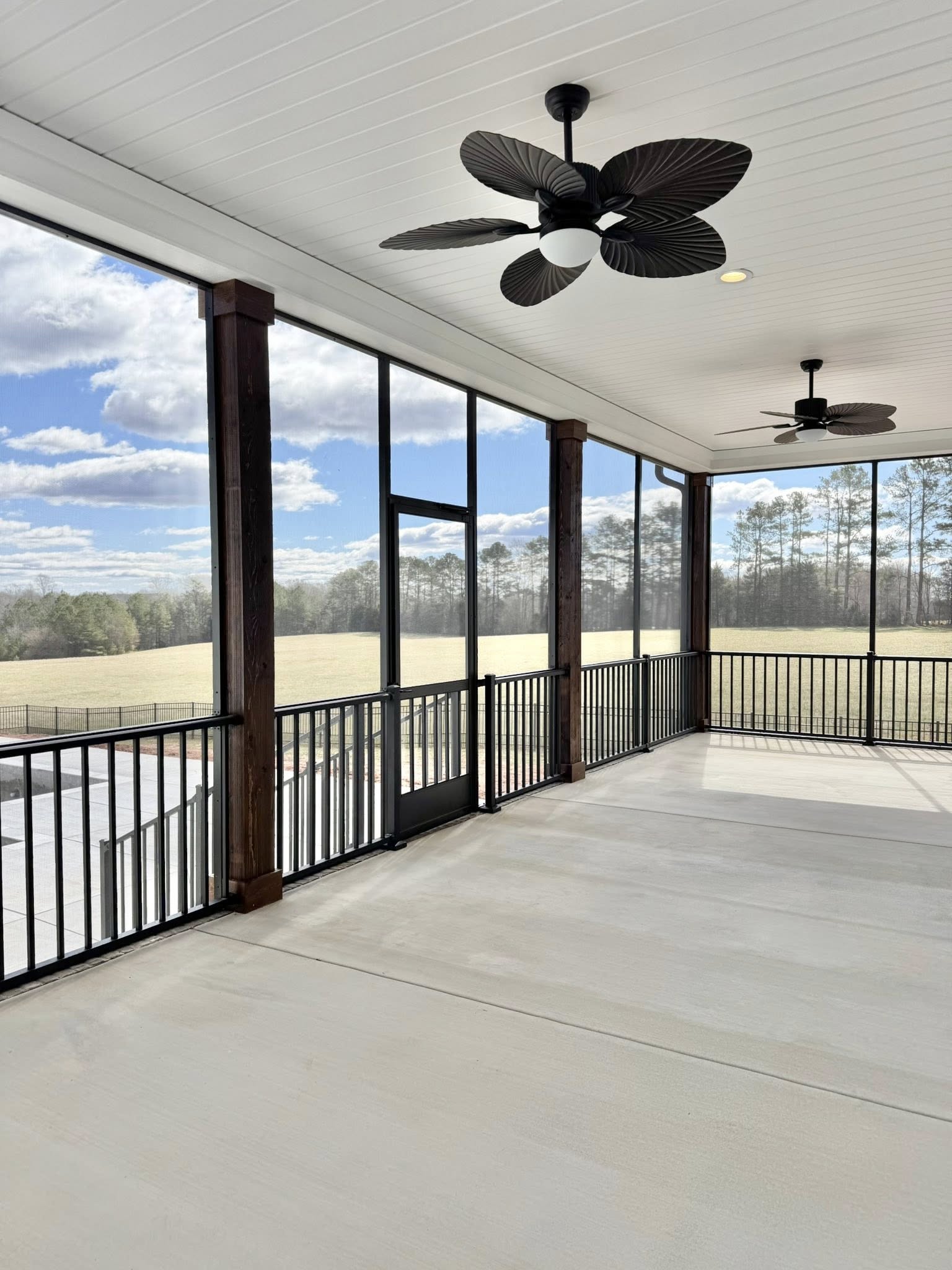 A spacious screened porch with two ceiling fans, wood columns, and black railings overlooks a sunny field and trees under a blue sky with clouds.