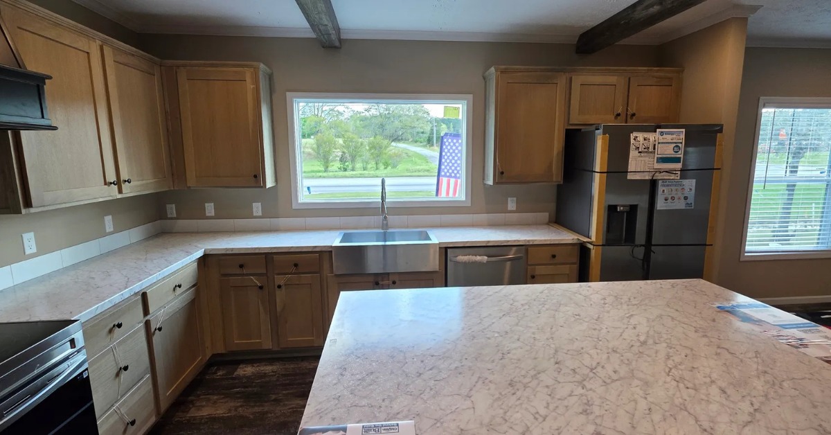 Spacious kitchen with light wooden cabinets, marble countertops, and a large center island. A window above the sink shows greenery outside. An American flag decorates the window.