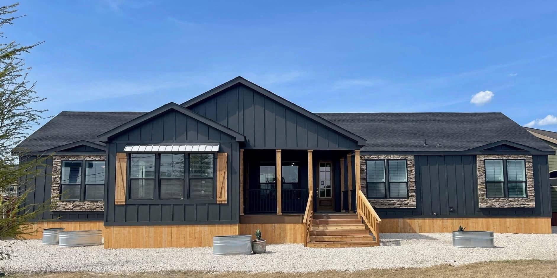 Alt text: Modern black and stone-clad house with a gabled roof, small porch, wooden steps, and planters. Bright blue sky, conveying a serene charm.