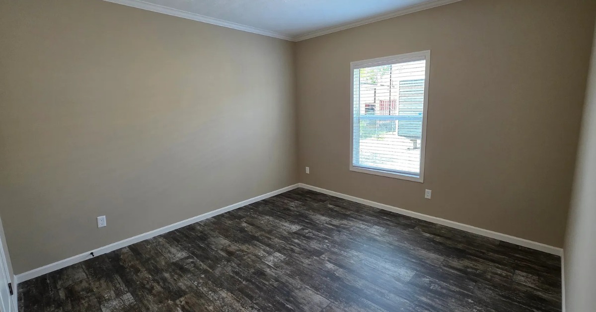 Empty room with beige walls, dark hardwood flooring, and a single window with blinds letting in natural light, creating a calm, spacious feel.