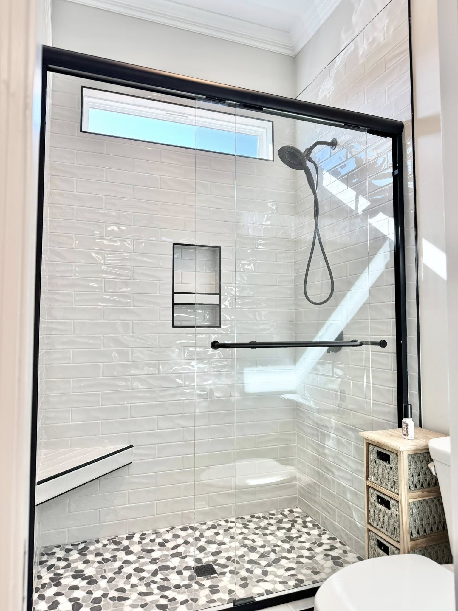 Bright modern shower with white subway tiles, black fixtures, pebble floor tiles, and a glass door. A built-in shelf and small window add elegance.