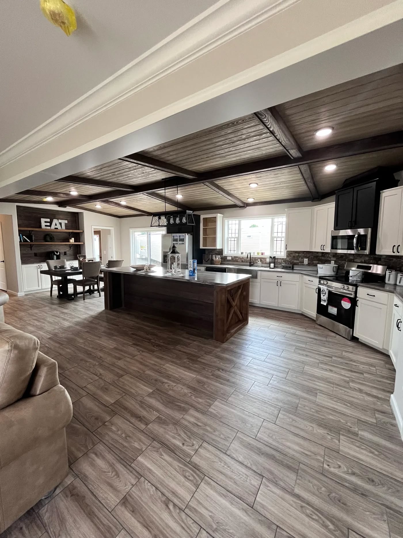 Spacious kitchen with wooden floors, large island, white cabinets, and black accents. Dining area visible with "EAT" sign. Cozy, welcoming vibe.
