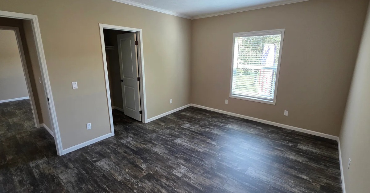 An empty room with beige walls and dark wood flooring. A window with blinds lets in natural light, and there's an open doorway leading to another room.