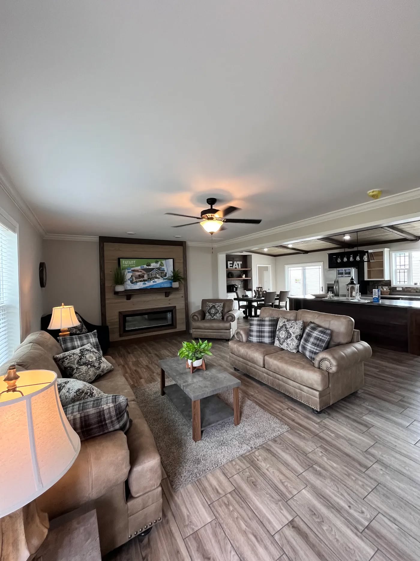 Spacious living room with brown leather sofas, plaid pillows, and a wooden coffee table. A TV and fireplace are on the wall. The kitchen is visible in the background.