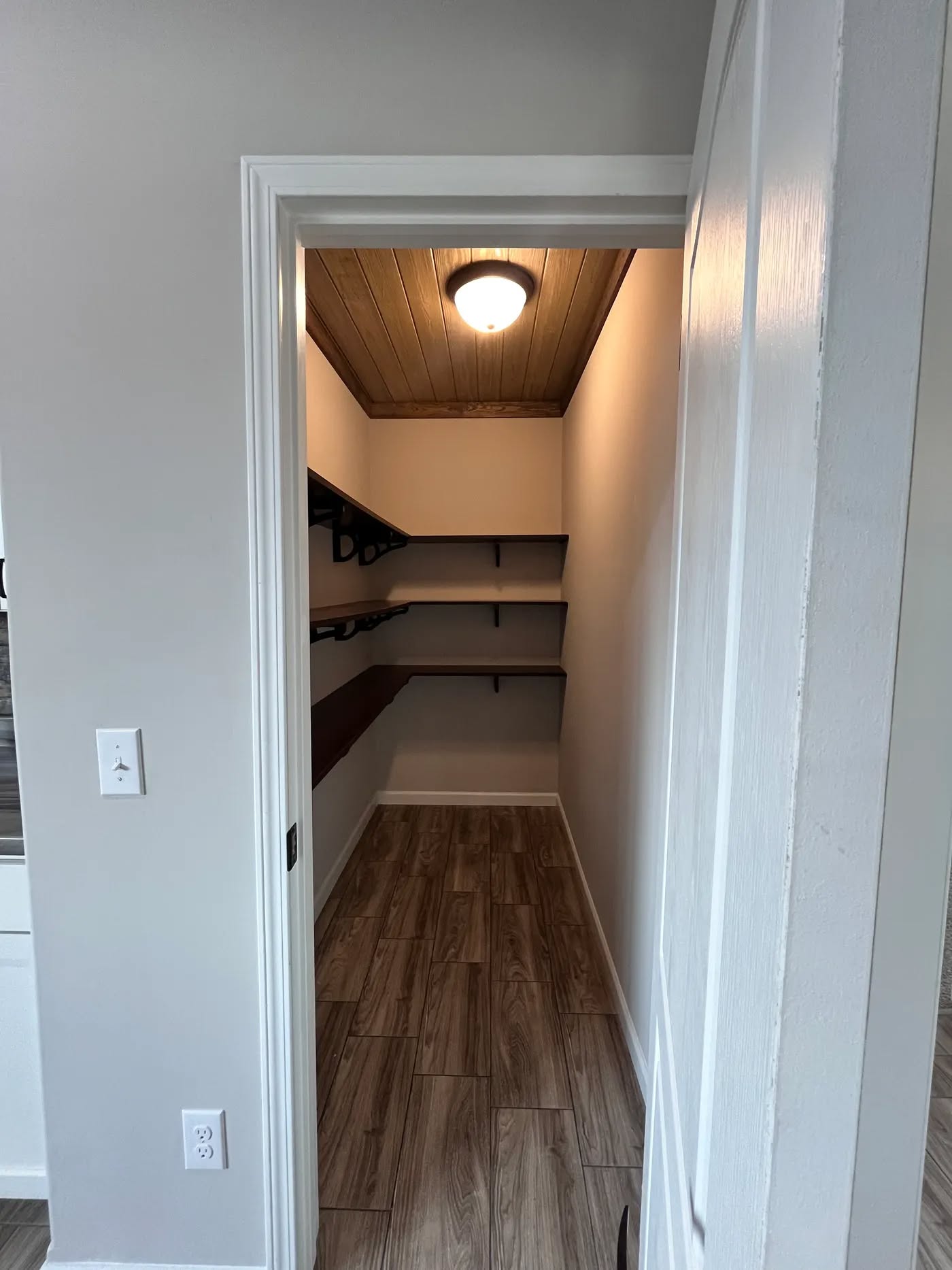 Narrow walk-in closet with a wooden ceiling, illuminated by a ceiling light. It has three dark wooden shelves on the left wall and light wood flooring. Neutral tones create a calm, organized atmosphere.