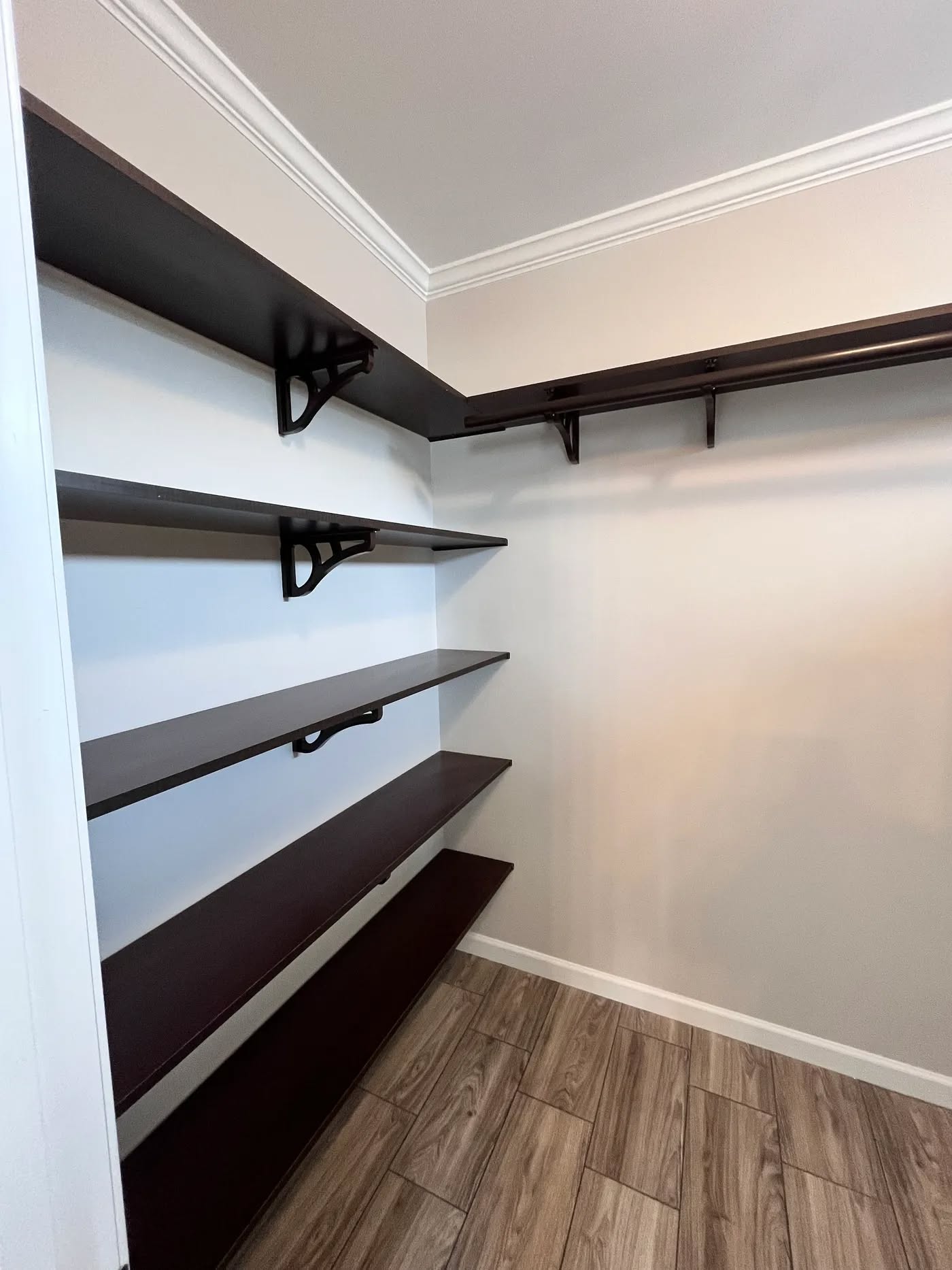 A corner closet features dark wooden shelves, metal brackets, and light gray walls, conveying a modern and organized feel. The floor is a light wood pattern.