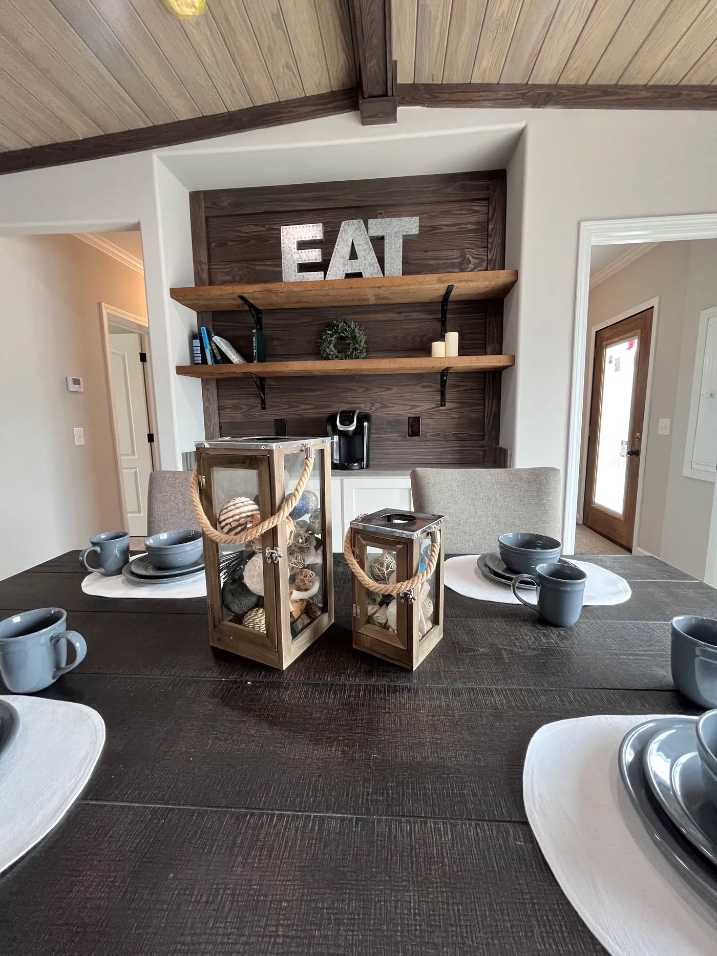 Dining area with a rustic table set for six with gray dishes and cups. Decorative lanterns with shells are centered. “EAT” sign on wood shelves.