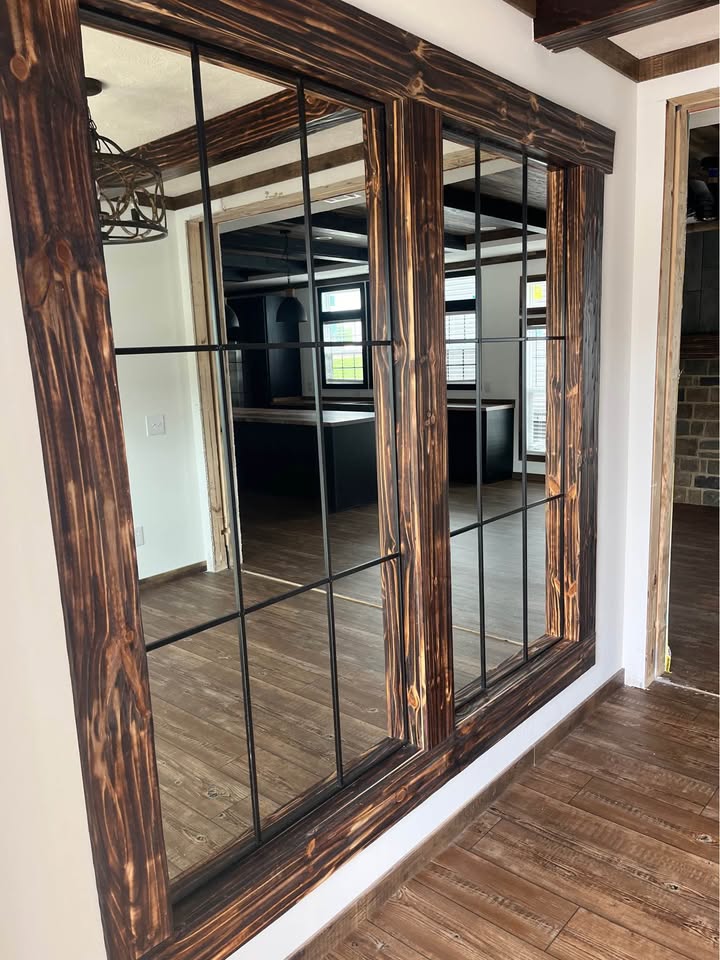Alt text: Large framed mirror with grid pattern reflects a wooden floor and modern room with beams and a window, creating a spacious and elegant feel.