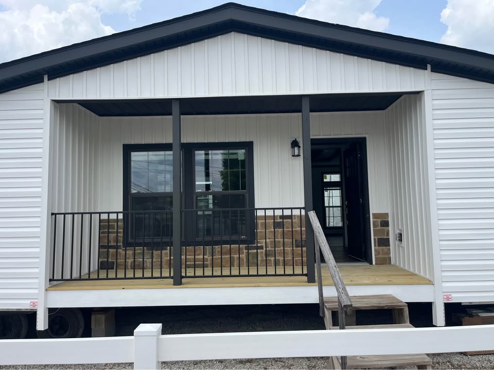 Small modular home with white siding and black trim. Features a covered porch with a black railing, wooden steps, and a stone accent wall.