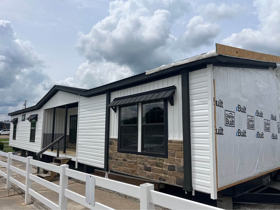 A partially completed modular home with white siding and stone accents sits beneath a cloudy sky. Wrapped in construction material, it exudes a work-in-progress feel.