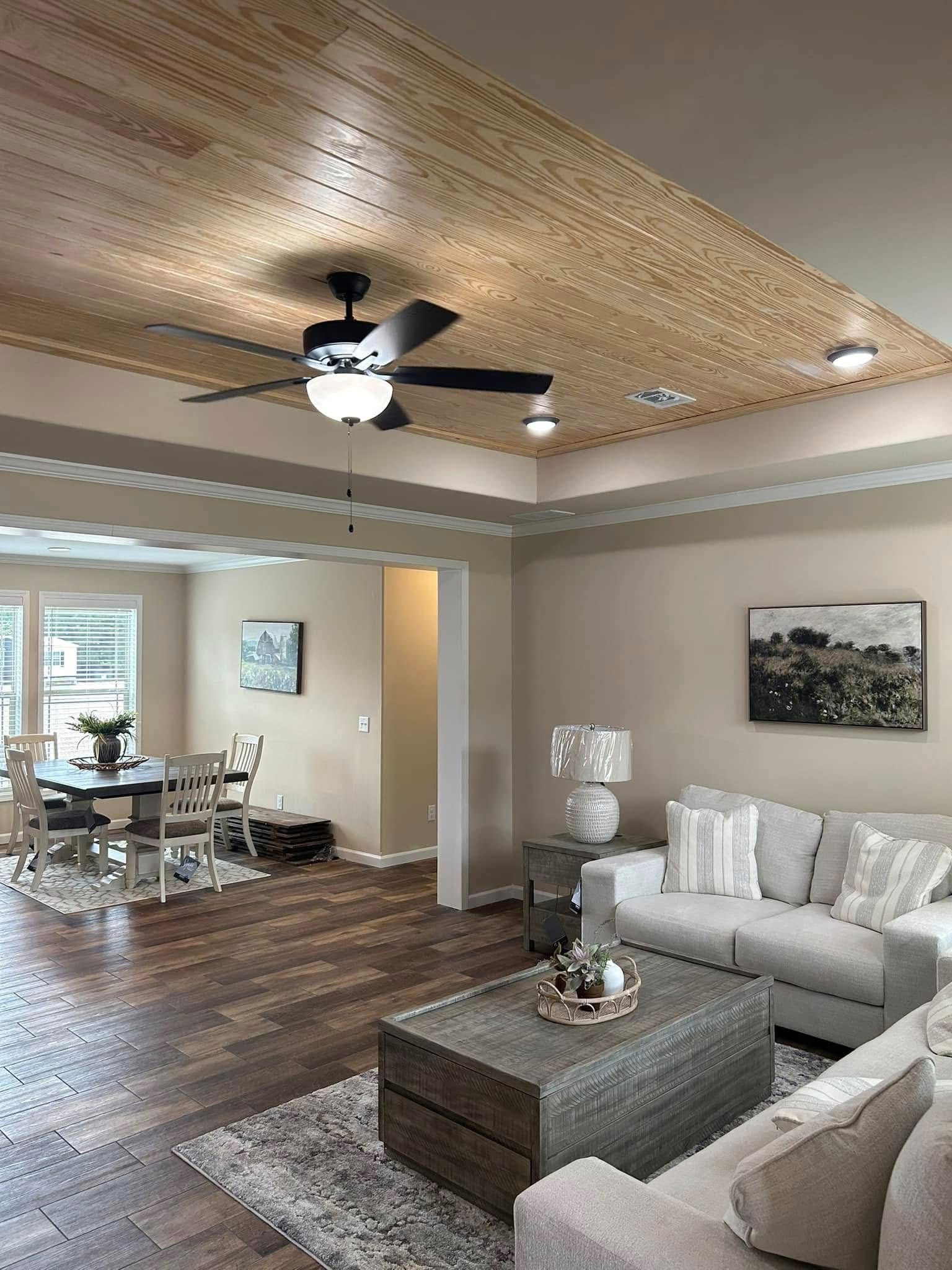 Modern living room with wood-paneled ceiling and fan. Cozy beige sofas, wooden coffee table, and lamp. Open dining area with windows in the background.