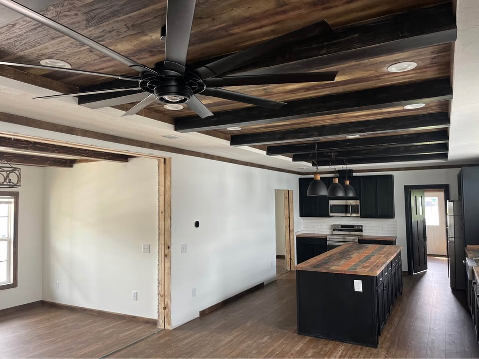 Modern kitchen with black cabinets and wooden countertops. Ceiling features rustic wooden beams and a large black fan. Bright, open layout.