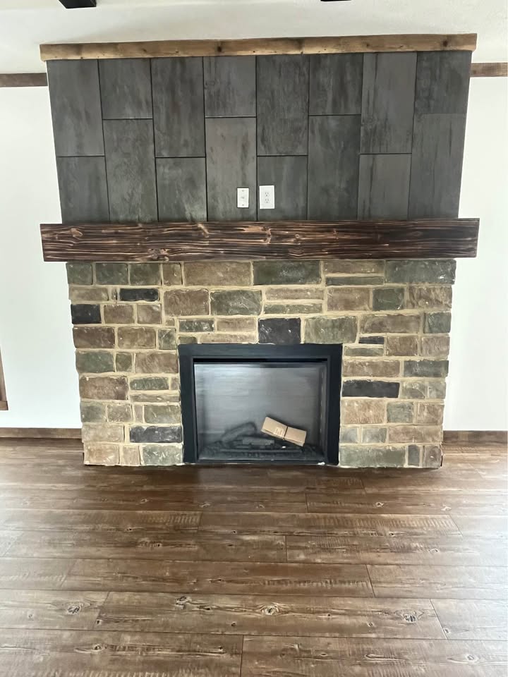 Stone fireplace with a dark wood mantel and textured gray tiles above. The floor is brown wood, and the room conveys a cozy, rustic feel.
