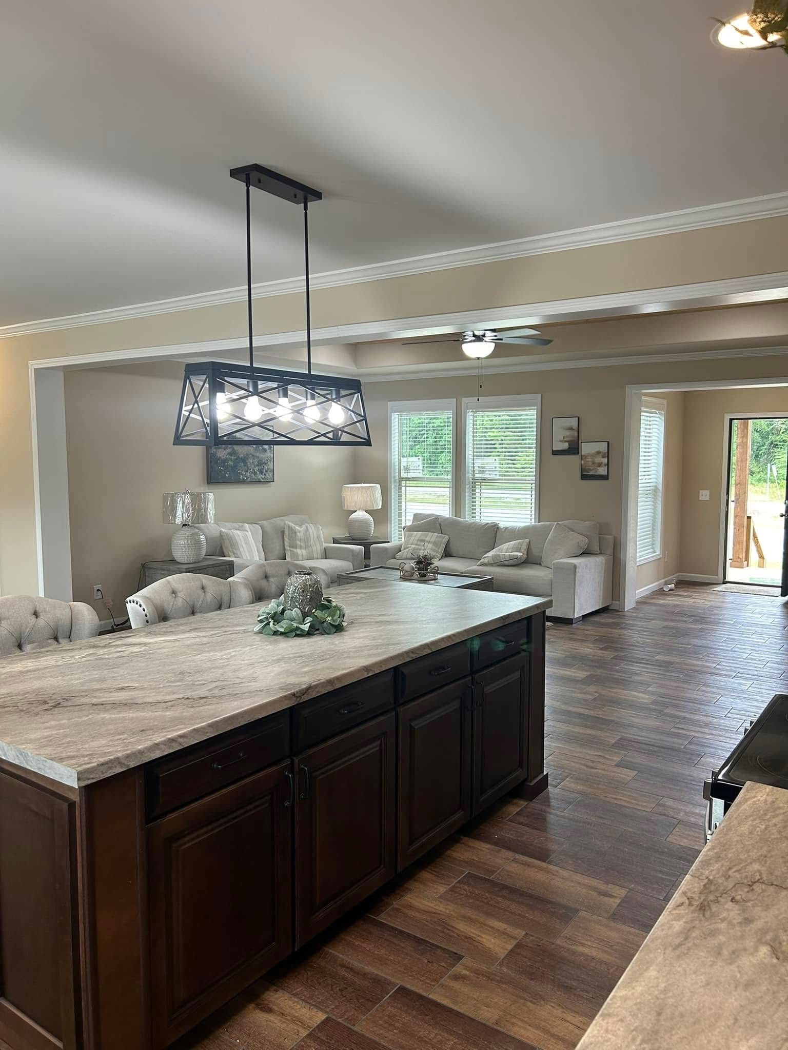 Spacious living room with a large kitchen island in the foreground. Modern chandelier overhead, cozy beige sofas, and large windows with lush greenery outside.