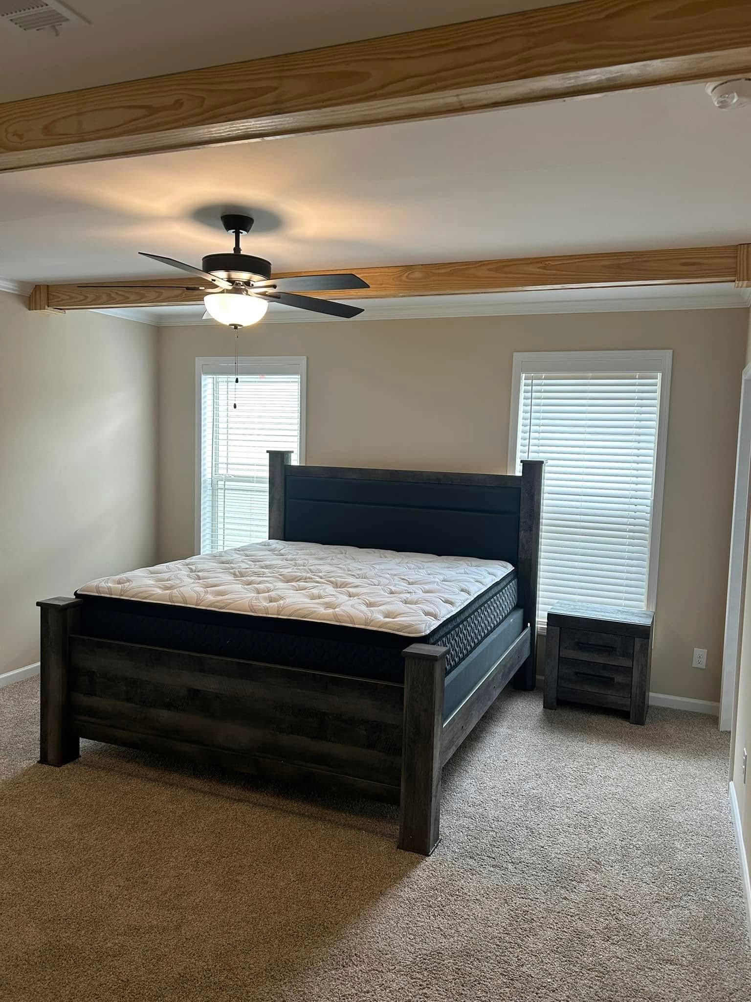 A cozy bedroom with a dark wooden bed frame features a neatly made mattress. Beige walls, neutral carpet, and dual windows create a serene ambiance.