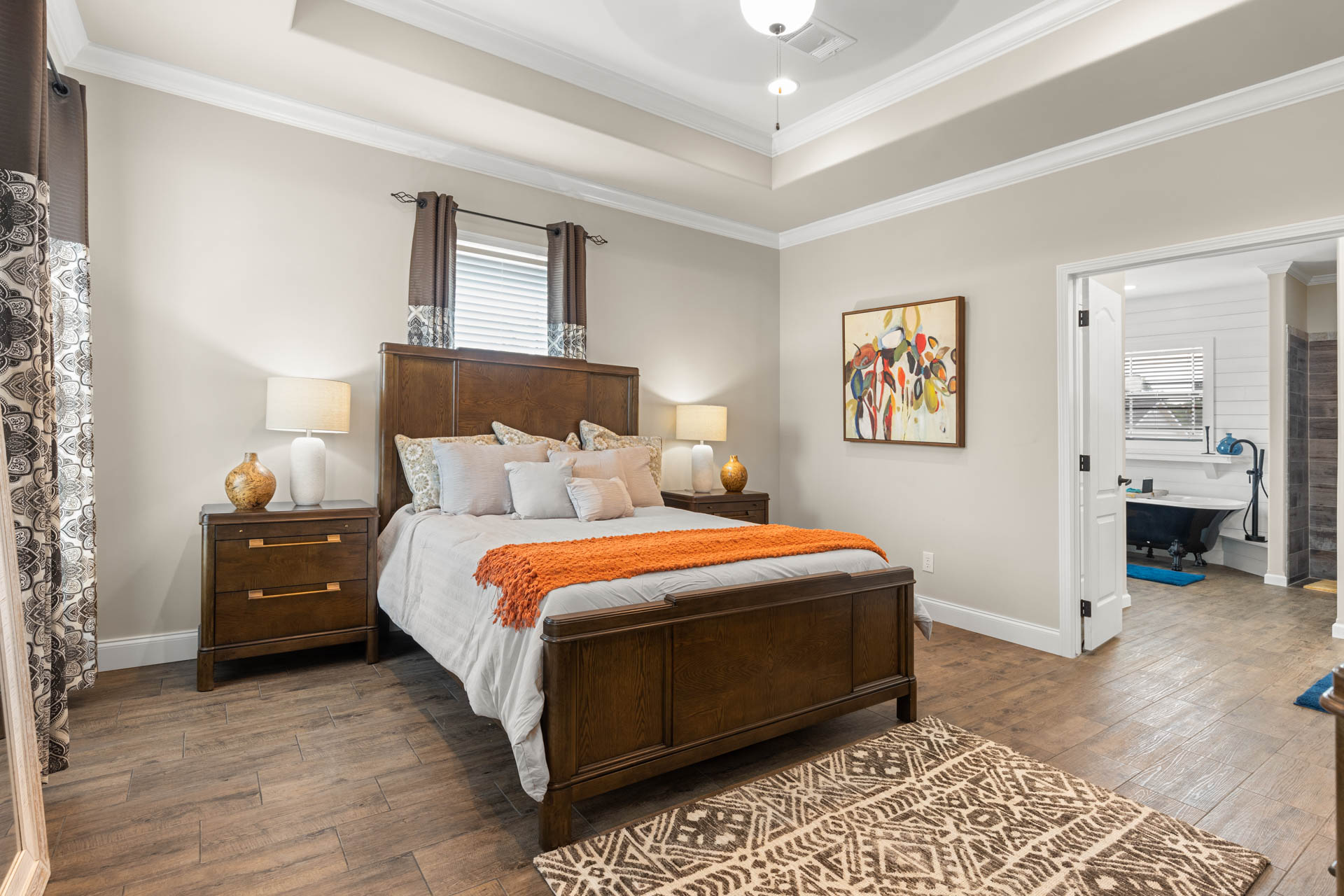 Modern bedroom with a wooden bed, orange throw, and patterned rug. Twin lamps on nightstands add warmth. Art hangs on a neutral wall, and a bathroom is visible.